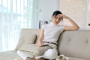 Obraz premium Young Asian woman sitting on sofa, holding hand to forehead, suffering from headache, feeling sick and stressed