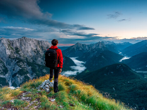 Hiker with backpack standing on a mountain ridge overlooking a misty valley at sunset for an adventure travel concept, Mangart, Julian Alps, Italy-Slovenia border.