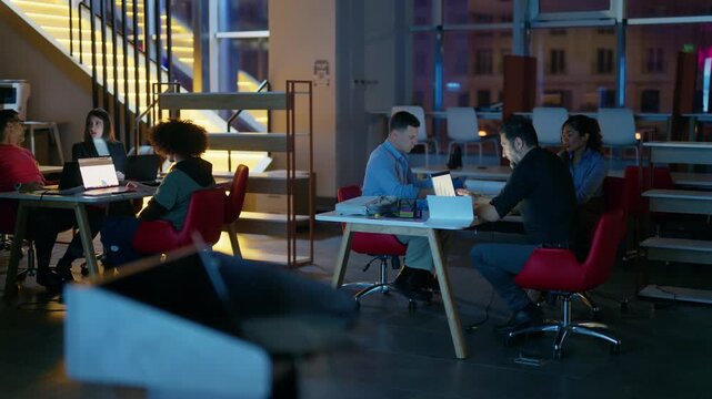 A diverse group of colleagues works late on their laptops in a modern office environment. They are collaborating and discussing projects during an evening work session.