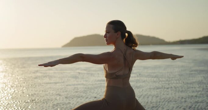 Portrait of athletic female stands in warrior asana on wet sand of sunrise ocean beach. Positive woman enjoys yoga exercising at exotic seaside. Self-care