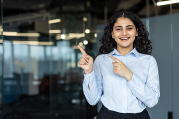 Smiling businesswoman in a light blue shirt pointing both index fingers to the left, indicating,...