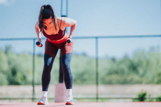 A fit woman performs bent-over dumbbell rows on an outdoor court. She focuses on strength training while wearing athletic workout clothes.