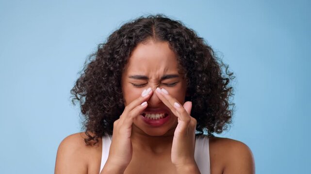 A woman with curly hair touches her face while expressing emotions. The background is blue, enhancing the focus on her feelings. The setting appears bright and well-lit.