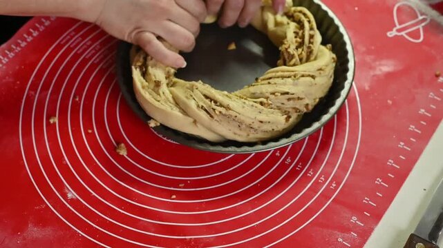 Dough wreath placed into round baking pan by hands, twisted pastry with nut filling arranged inside mold, preparation before baking, closeup kitchen scene, homemade dessert concept,