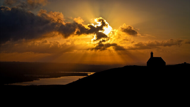 Aerial view of Chapelle Saint Michel de Brasparts silhouetted on a hill with dramatic sun rays piercing through golden clouds over a lake Saint-Rivoal, Bretagne, France.