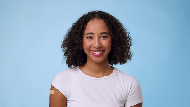 Young African American woman with curly hair smiles confidently after getting a vaccination. She has a bandage on her arm and stands in front of a light blue background.