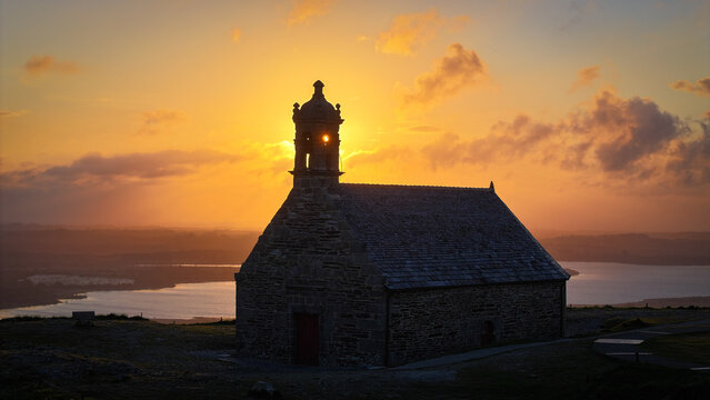 Aerial view of Chapelle Saint Michel de Brasparts silhouetted against a golden sunset with the sun glowing through the bell tower in Saint-Rivoal, Bretagne, France.