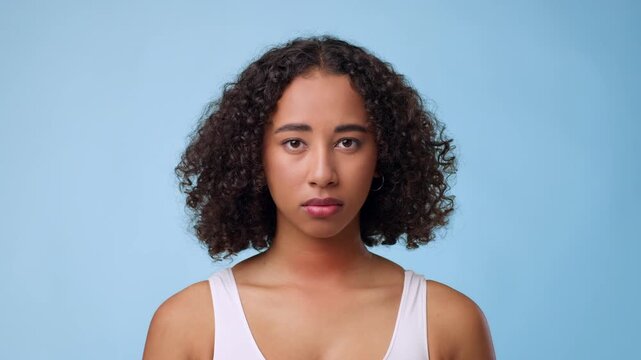 A young woman with curly hair stands confidently against a light blue background. She looks directly at the camera with a serious expression. The studio lighting highlights her features.