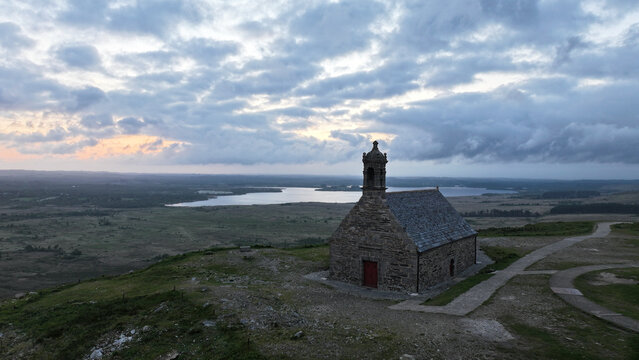 Aerial view of Chapelle Saint Michel de Brasparts on a hilltop overlooking the Lac de Saint-Michel under a cloudy sky at dusk in Saint-Rivoal, Bretagne, France.