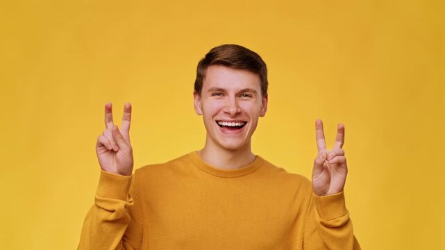 A young man with light skin and brown hair smiles widely while making peace signs with both hands. He stands in front of a vibrant yellow backdrop, showcasing joy and positivity.