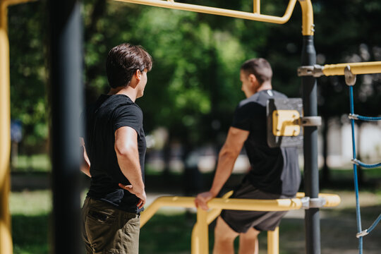Two men train at an outdoor fitness station in a leafy park. One man watches while the other uses parallel bars for a bodyweight exercise.