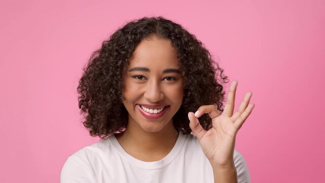 A young woman with curly hair is smiling and showing an OK hand sign against a pink background. She conveys happiness and positivity.