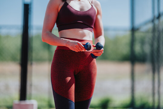 A young woman wearing a sports bra and leggings holds small hand weights while preparing to exercise. She stands outdoors by a fence ready for a strength workout in casual athletic wear.