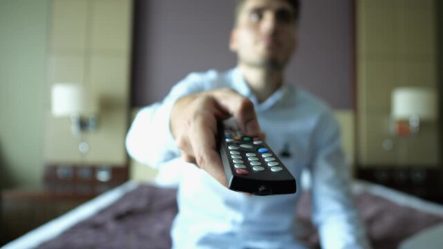 Focus on male hand of young businessman sit on bed switch channels on TV remote control at modern hotel room. Cozy relaxation during business trip showing comfort and leisure with blur background
