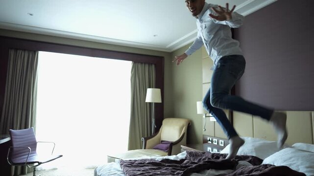 Happy young businessman jumping on bed in modern hotel room with joyful emotion and carefree mood. Positive man enjoying comfort and freedom during business trip showing travel experience indoor