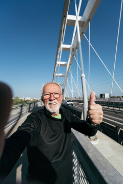 Active senior man in sportswear taking a selfie video or vlogging on a modern bridge, smiling at the camera and showing a thumbs up gesture during his outdoor running routine.