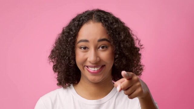 A woman with curly hair smiles and points directly at the camera. She stands in front of a solid pink background. The setting appears bright and engaging, conveying a friendly atmosphere.