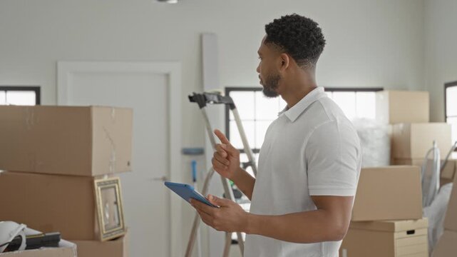 Man holding blue tablet and pointing finger amid stacked moving boxes, ladder and packing materials in apartment; focused planning.