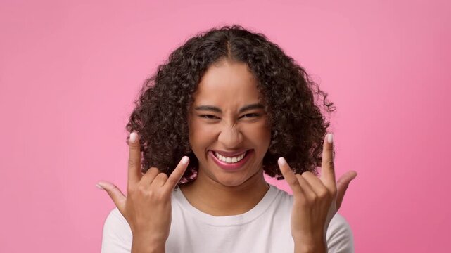 A young woman with curly hair makes a rock gesture with her hands. She smiles widely against a pink background. The scene captures her joyful expression and energy.