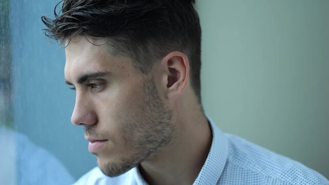 Portrait of sad young entrepreneur sitting near window in modern hotel room or office. Unhappy man with serious expression looking away showing emotional stress, fatigue and professional burnout