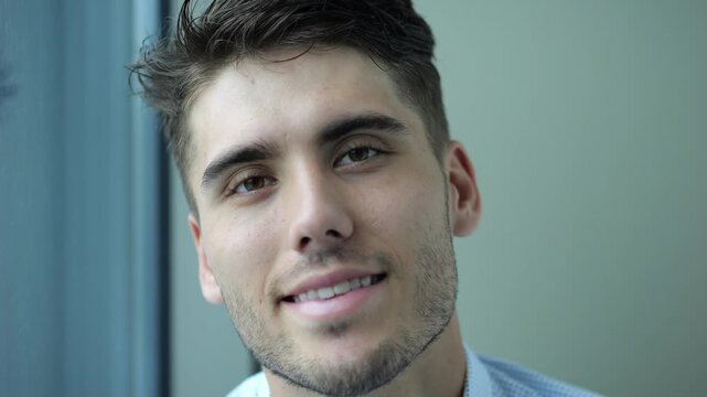 Portrait of happy businessman sitting near window in modern hotel room or office. Relaxed young manager looks into camera with smile showing relief, confidence and success in professional lifestyle