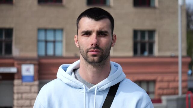 Portrait of young confident bearded man in casual clothing with cityscape background. Close up of handsome guy looks into camera standing at city environment at sunny day