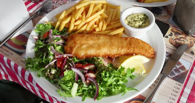 Traditional fish and chips plate served with crispy golden french fries and tartar sauce bowl. Deep fried battered cod fillet sits alongside fresh green salad mix and lemon wedge on white ceramic dish