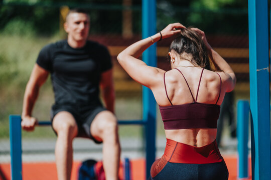 A fit woman adjusts her hair while standing at outdoor parallel bars during a workout. A man trains in the blurred background as the pair exercise at an outdoor gym.