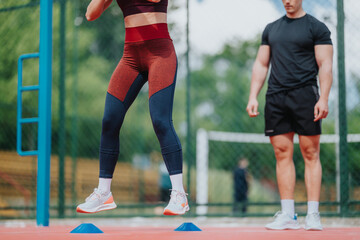 Fototapeta premium Female athlete performs a cone jump agility drill outdoors on a fenced court under trainer supervision. The trainer watches as she practices explosive footwork and jumping between markers.
