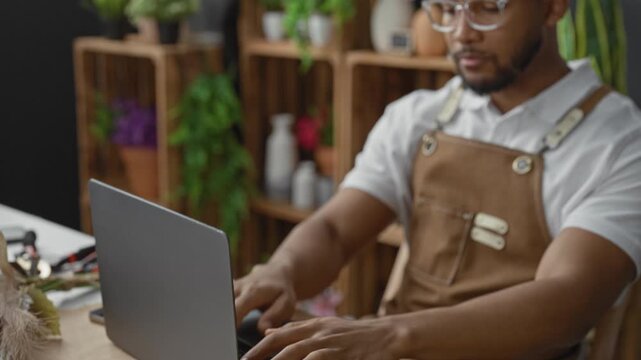 Man florist typing on laptop in studio, wearing apron and glasses, surrounded by plants and vases; focused productivity planning.