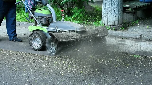 Worker operating small road cleaning machine, green vehicle with rotating brush clearing dirt and debris from roadside, outdoor environment with grass and trees, road maintenance in process
