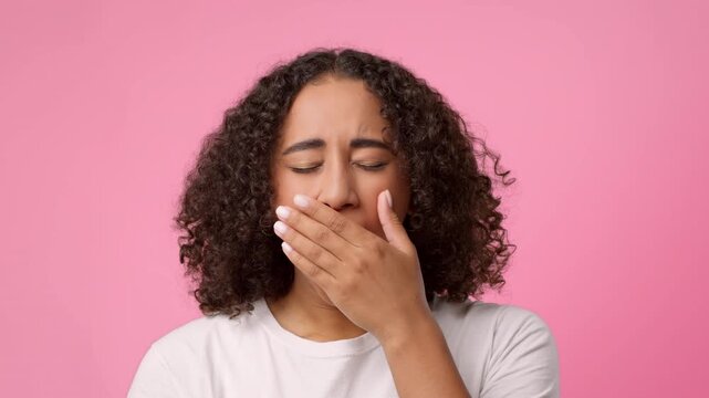 A woman with curly hair covers her mouth while yawning. She appears tired or bored in front of a bright pink background. This moment captures a relatable expression of fatigue.