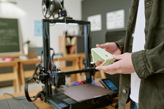 Anonymous student holding 3D printed prototype beside printer in workshop, demonstrating product design and rapid manufacturing