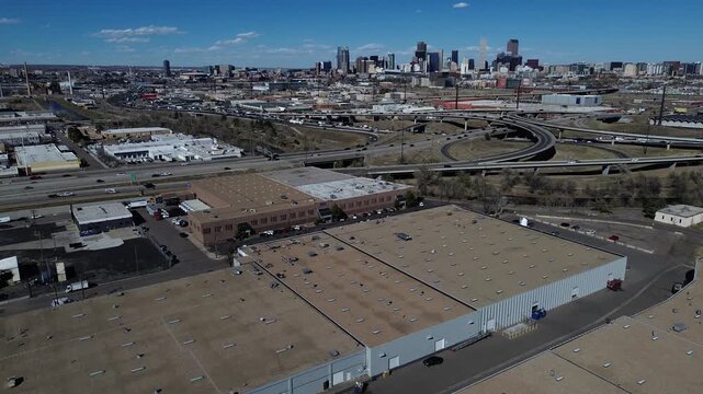 Skyline interchange span showing warehouse blocks near W 4th Ave with layered highway ramps leading toward downtown Denver. Flat roofs, rooftop vents HVAC, paved lots, logistic distribution, CO