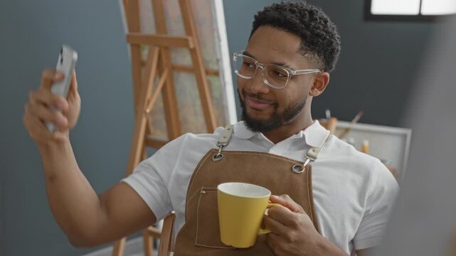 Man holds smartphone and yellow mug, wearing apron and glasses, smiling while checking phone in studio with easel and paint brushes; contentment creativity.
