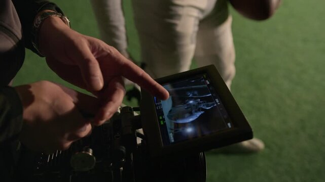 Close-up of hands of male cinematographer reviewing footage of unrecognizable gridiron player with helmet walking on pitch, using professional video camera, while filming for documentary or commercial