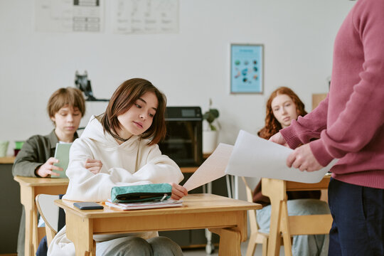 Teenage girl receiving test paper from teacher while sitting in classroom with classmates, showing school assessment and academic performance