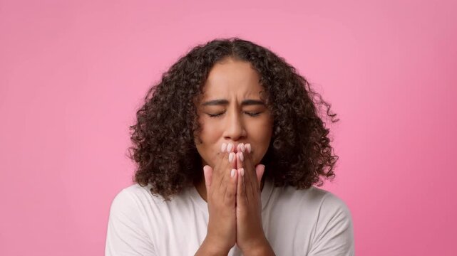A woman with curly hair stands in front of a pink background. She has her hands on her face and looks surprised. This moment captures a relatable expression.