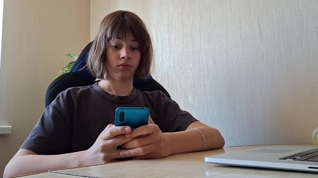 Teen sits at desk and uses phone while looking focused in quiet indoor space