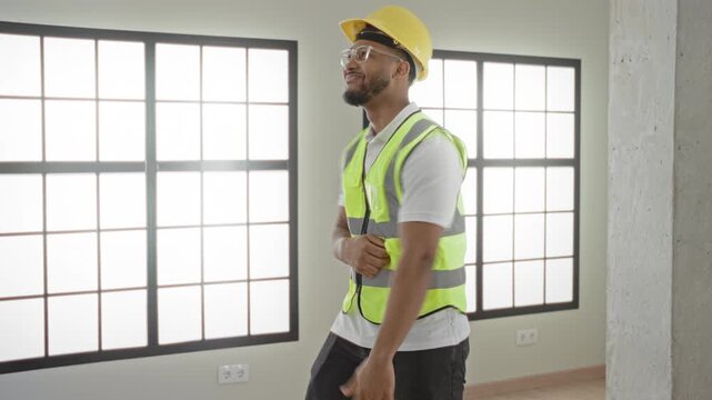 Man in hardhat and safety vest dancing with fists raised near large grid windows inside a bright unfinished building; celebration joy.