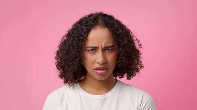 A young African American woman with curly hair shows a confused expression in front of a plain pink background. She wears a white shirt and has an inquisitive look on her face.