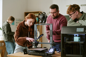 Teenagers and middle aged male teacher operating 3D printers during classroom workshop, demonstrating STEM prototyping and innovation © AnnaStills