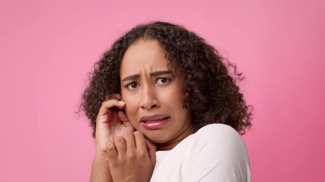 A young African American woman with curly hair displays a fearful expression while standing in front of a pink background. She is holding her hands near her face, showing surprise or concern.