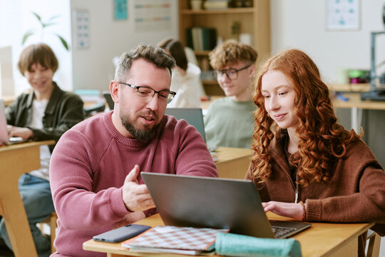 Middle aged Caucasian male teacher guiding teenage girl using laptop in classroom, supporting digital learning and mentoring