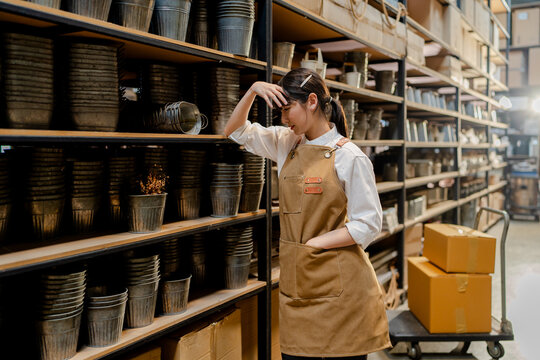 exhausted asian warehouse worker feeling sick headache stressed work in store rack, woman staff in apron working in distribution shipping logistic center support fulfillment delivery supply chain