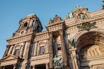 Obraz premium A detailed low-angle architectural photograph capturing the ornate facade, green domes, and intricate stone sculptures of the Berlin Cathedral in Germany under a clear blue sky.