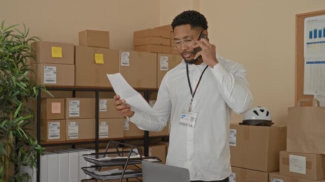 Young man on phone holding a document and reading labels while wearing a lanyard badge in a warehouse office; logistics focused.