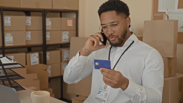 Man examining creditcard while speaking on smartphone to ear, holding card near face and scanning parcel labels in a storage building; concentration.