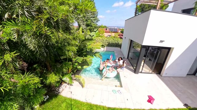 Upward aerial shot of a diverse group of happy friends having fun with inflatable toys in a swimming pool, enjoying their summer vacation at a modern luxury villa on a sunny day