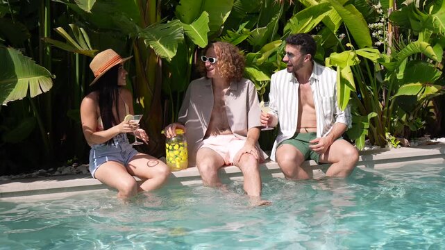 Group of cheerful young friends enjoying a tropical holiday, sitting on the poolside with their feet in the water, toasting with refreshing cocktails, talking and laughing together